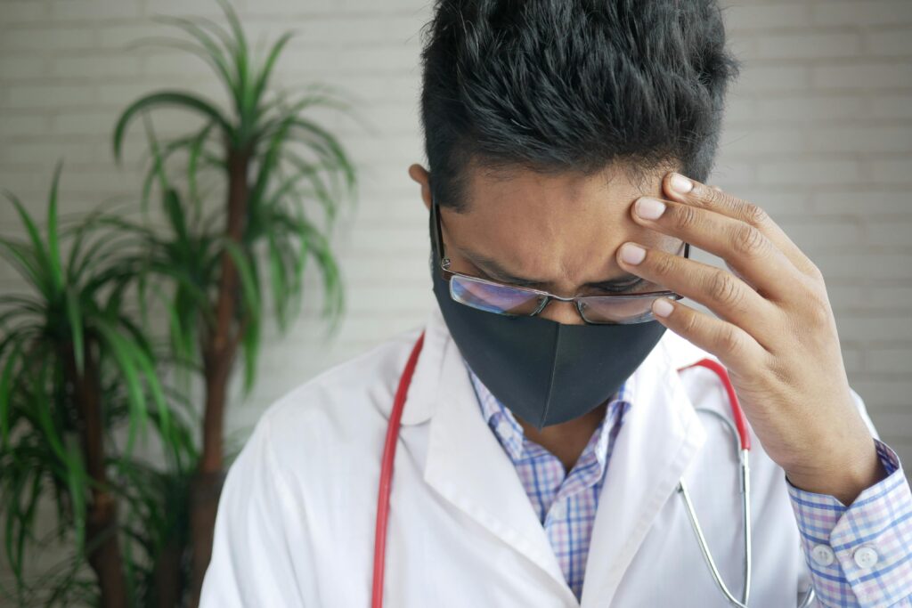 A thoughtful doctor wearing eyeglasses and mask indoors, appearing concerned.